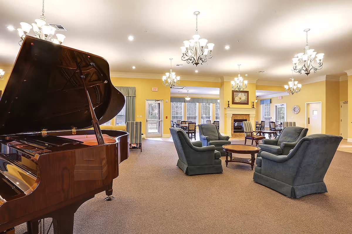 Spacious and well-lit common area with a grand piano on the left, several blue upholstered armchairs arranged around a wooden coffee table, and multiple chandeliers hanging from the ceiling. In the background, there is a fireplace with a framed flower painting above it and dining tables near large windows letting in natural light.