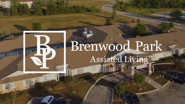 Aerial view of Brenwood Park Assisted Living facility showing a large single-story building with a brown roof, surrounded by parking spaces, trees, and open grassy areas. A curved driveway leads to the main entrance of the building.