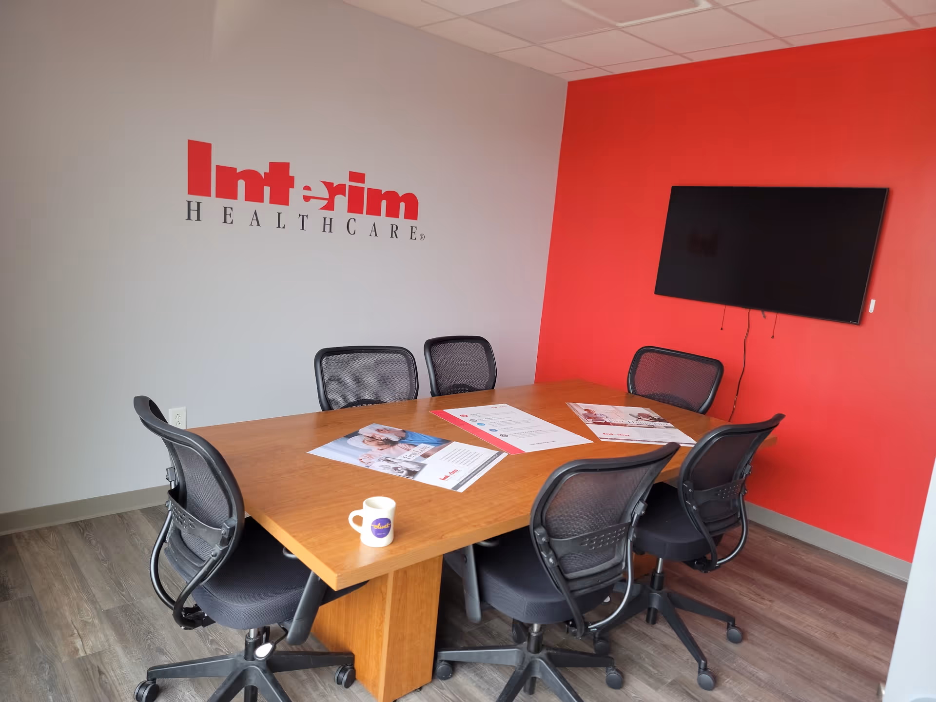 Conference room with wooden table, six chairs, and a mounted TV on a red accent wall.