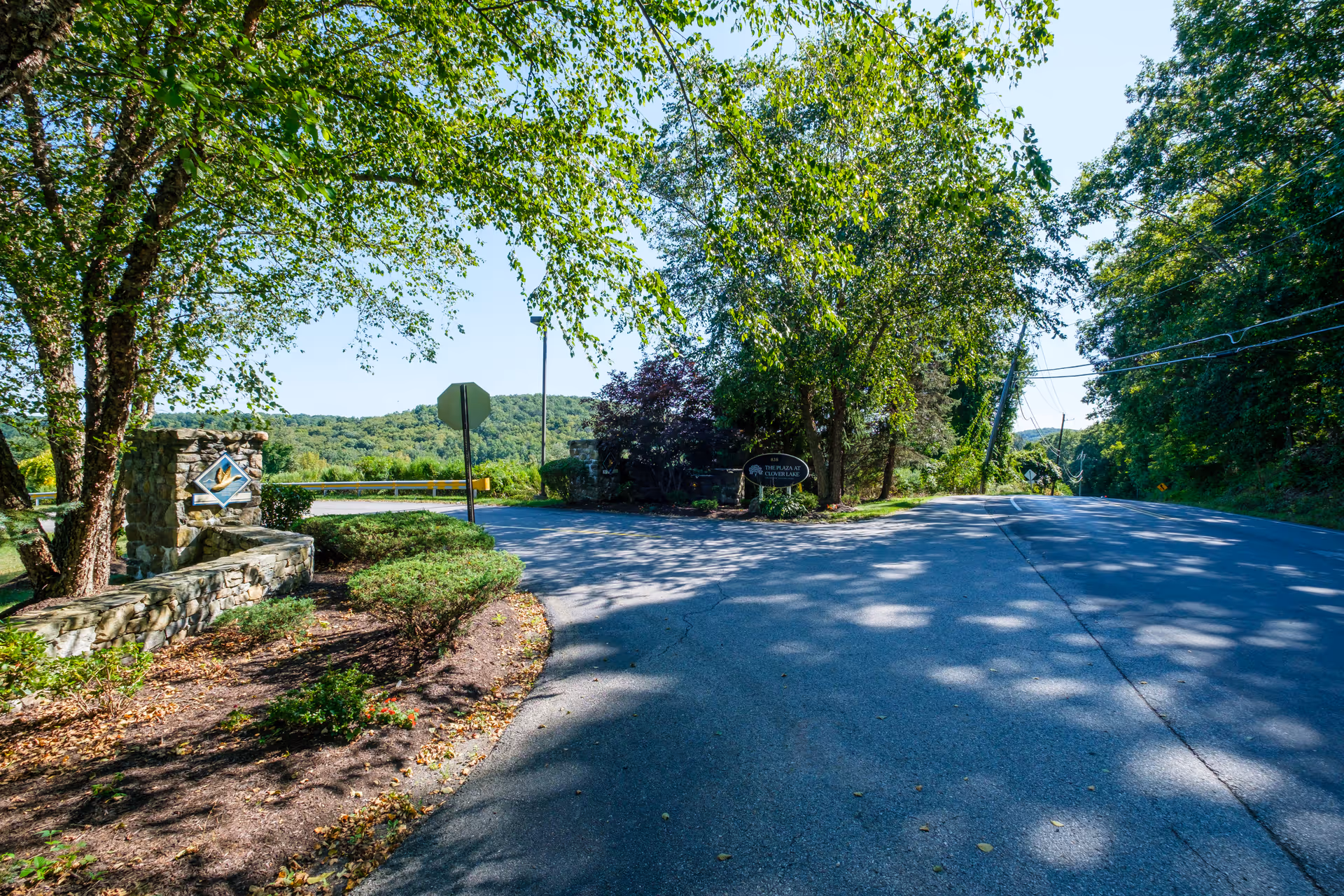 Paved road and landscaped stone entrance with a sign for The Plaza at Clover Lake under leafy trees.