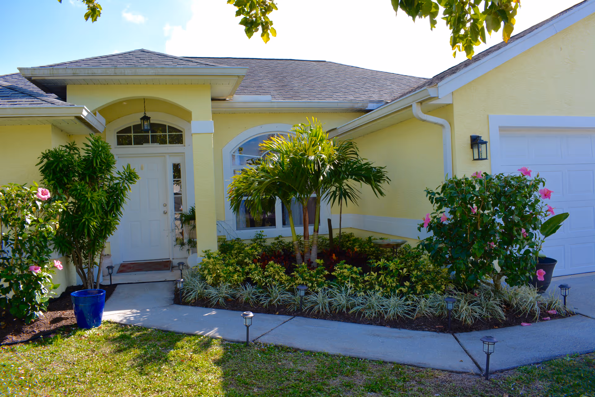Front exterior of a yellow senior living facility with tropical plants and a curved walkway