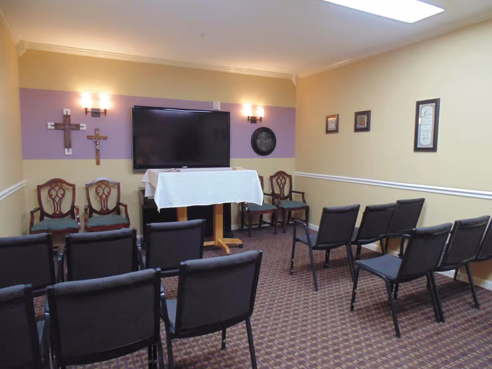 A small room set up for group gatherings with rows of black chairs facing a TV mounted on a wall with a purple and beige color scheme. There are four wooden chairs with green cushions along the walls, two crosses on the left wall, a clock on the right wall, and three framed pictures on the far right wall. A table covered with a white cloth stands in front of the TV.