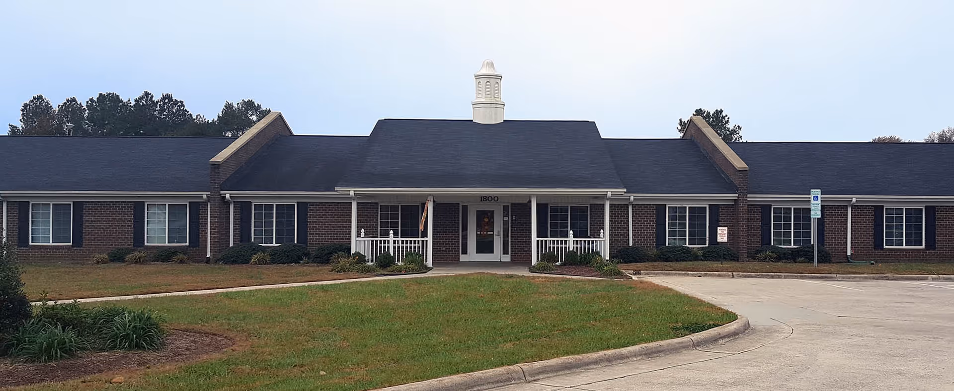 Single-story brick senior living building with a central entrance and cupola, surrounded by lawn and a parking area.