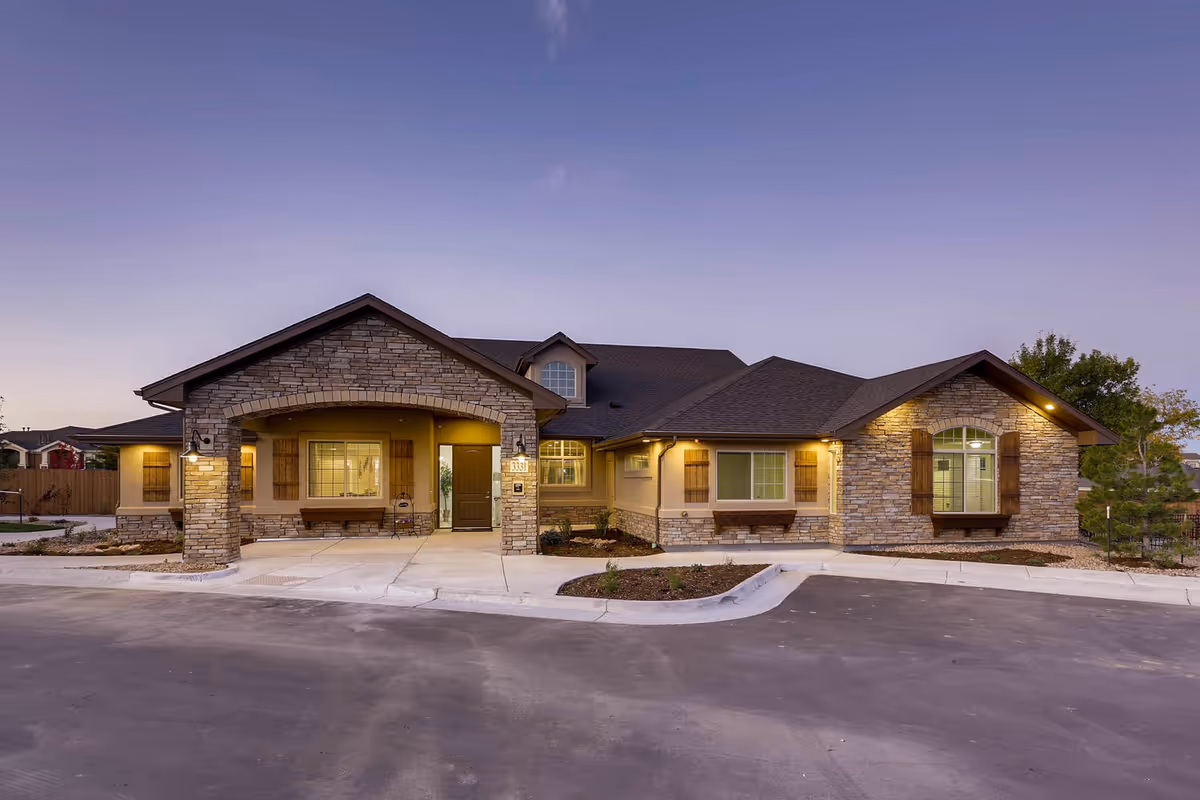 Single-story stone-faced senior living facility exterior with a welcoming covered entrance and warm lights.