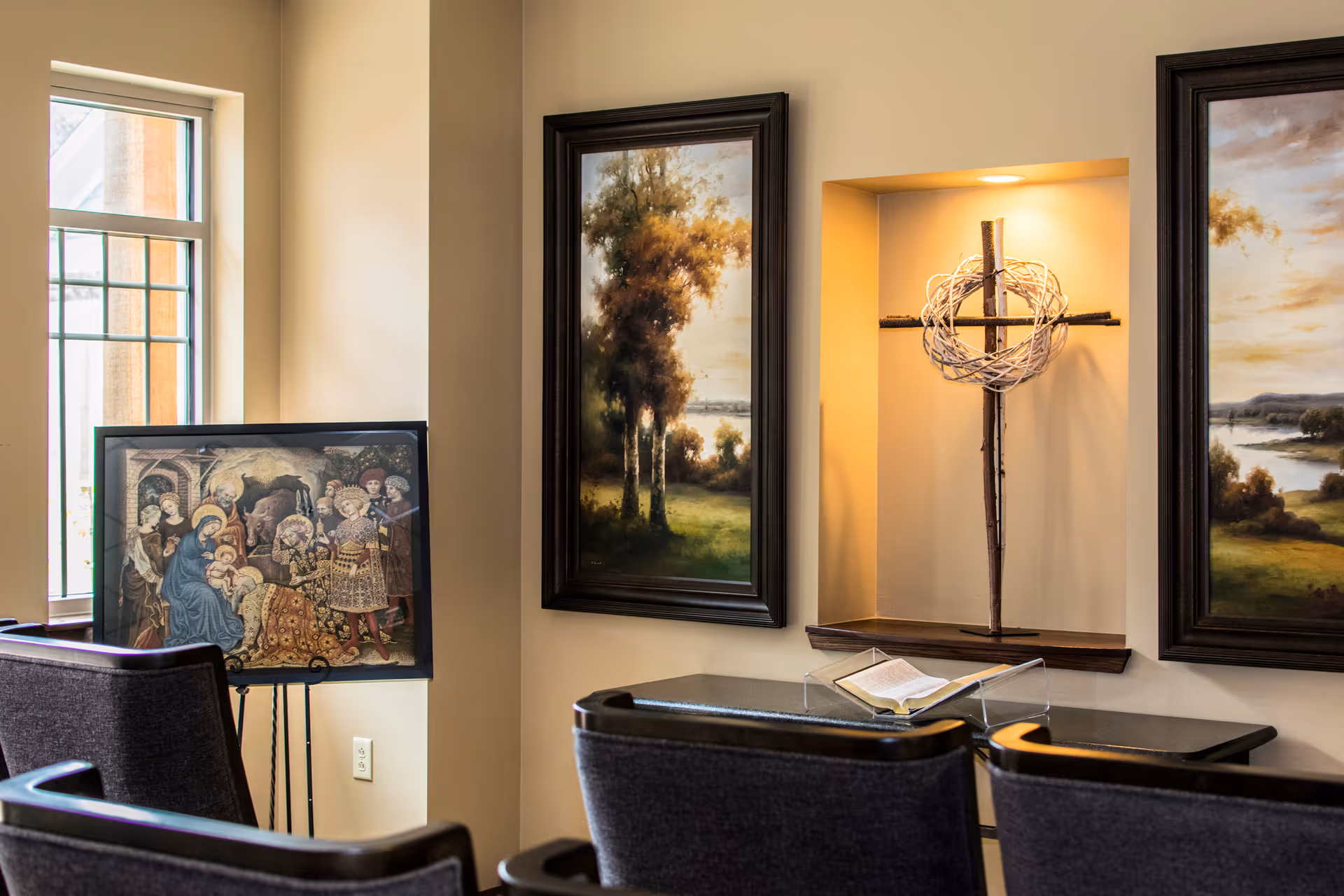 Interior room with framed paintings, a cross sculpture, and empty row of chairs.