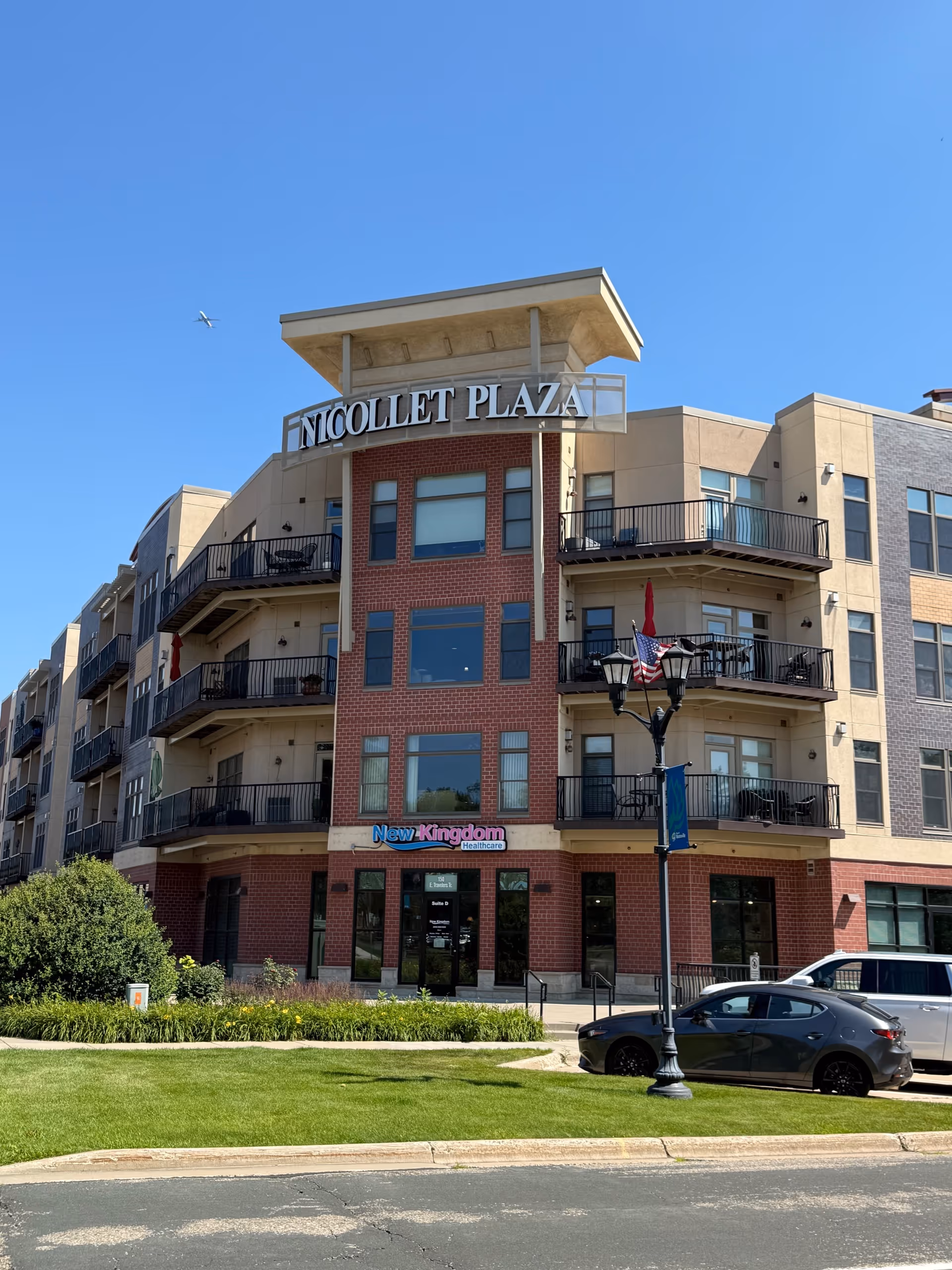 Three-story brick and beige senior living building with balconies, signage, and manicured lawn.