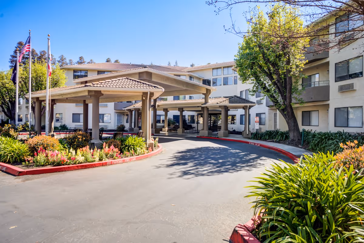 Entrance area of a senior living facility with a covered drop-off zone, surrounded by well-maintained landscaping including bushes and flowers. The building is multi-story with numerous windows, and there are three flagpoles with flags near the entrance.