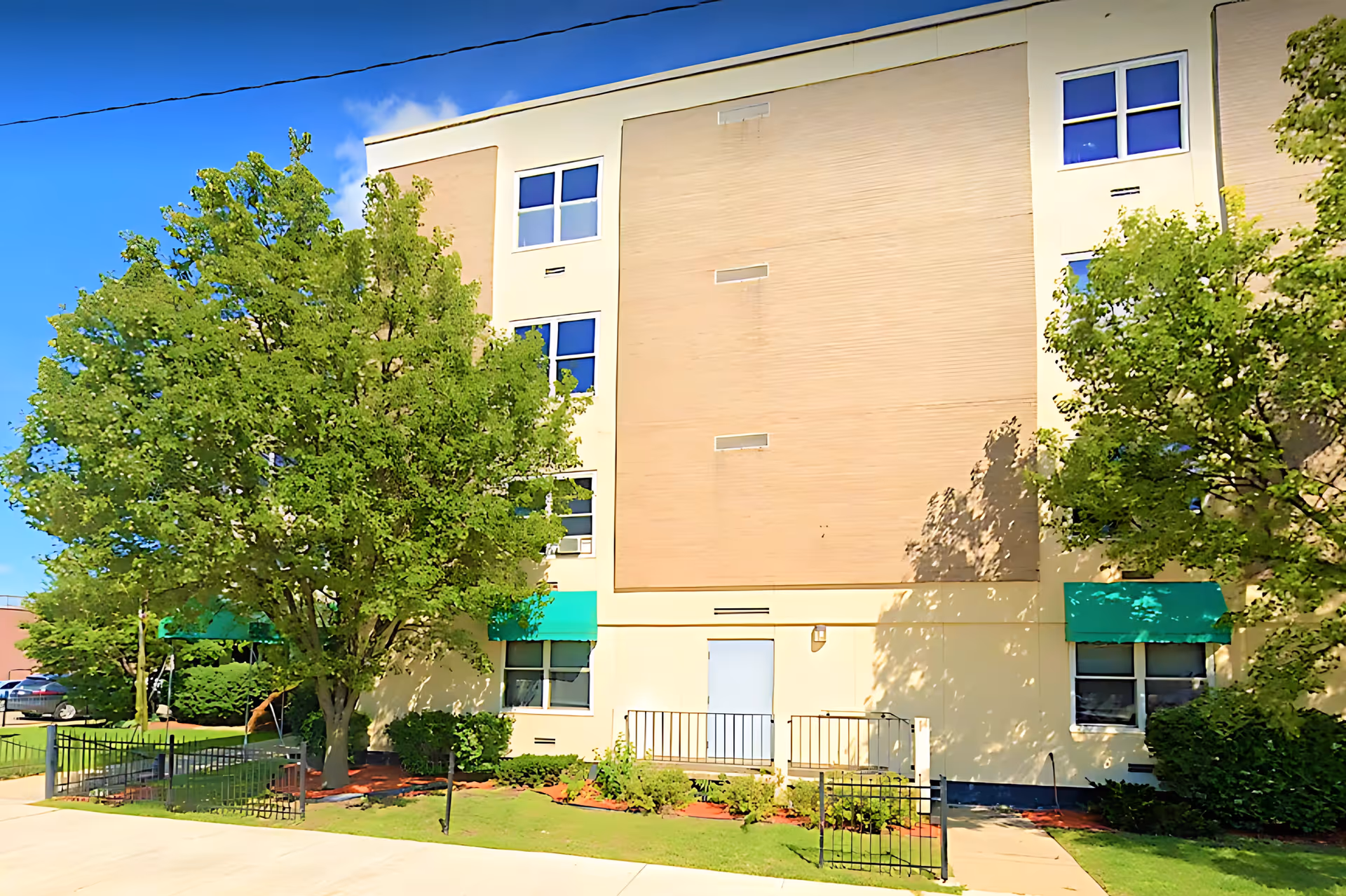 Exterior view of a multi-story building with beige and cream walls, several windows, green awnings, and trees in front. The building is surrounded by a small fenced garden area with shrubs and a sidewalk in front.
