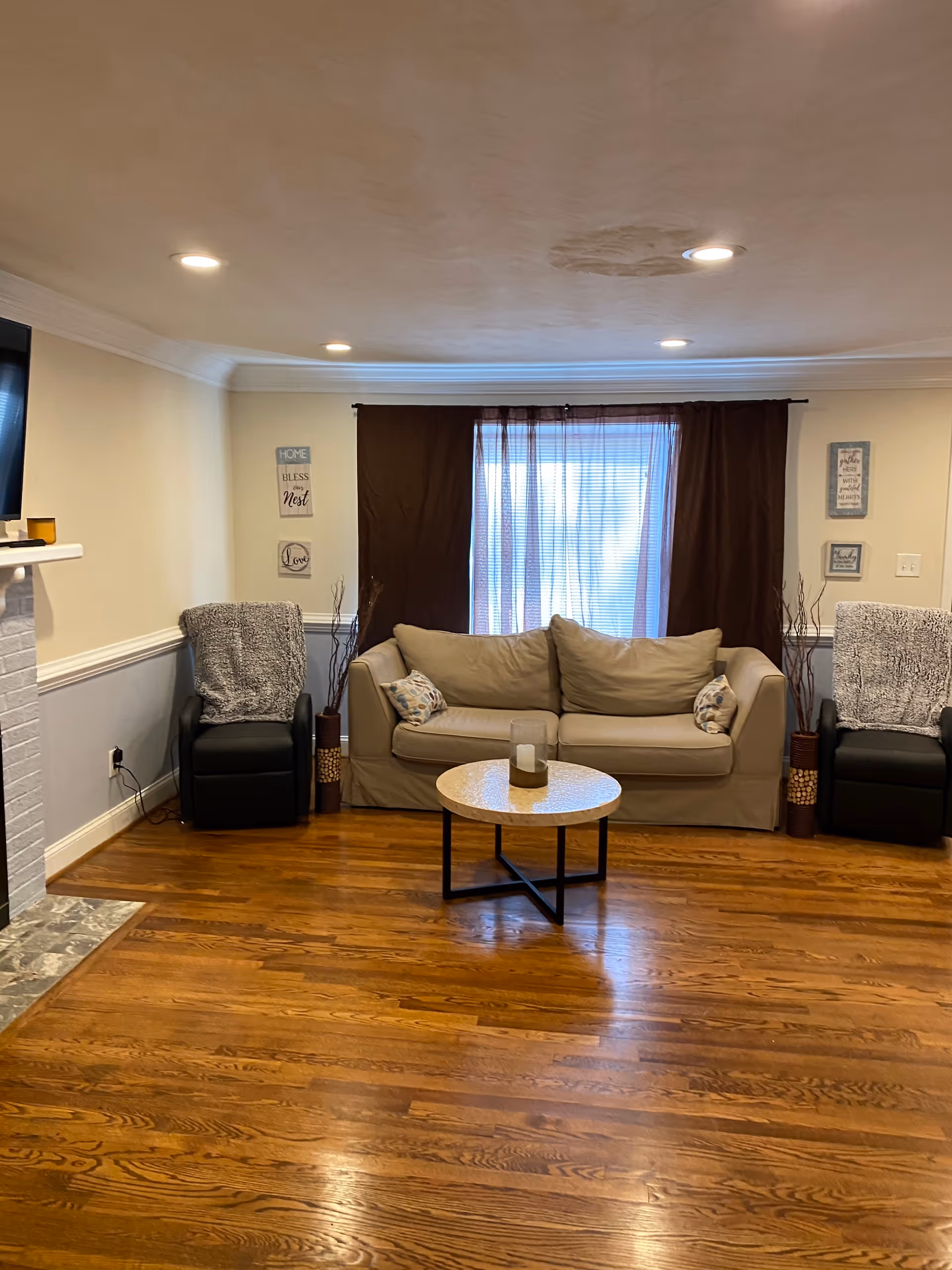 A cozy living room with a beige sofa flanked by two black armchairs covered with gray throws. A round coffee table with a candle centerpiece sits on a wooden floor. Behind the sofa is a window with brown curtains and sheer blinds. The walls are light-colored with decorative wall art and a white fireplace is partially visible on the left.