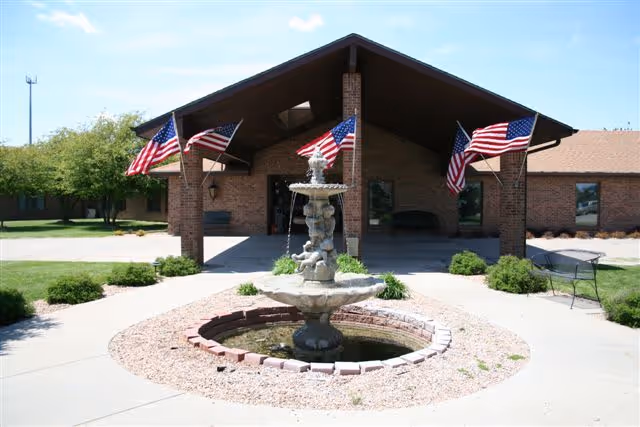 Front entrance of a brick retirement facility with a central fountain, landscaped beds, and American flags on the covered porch.