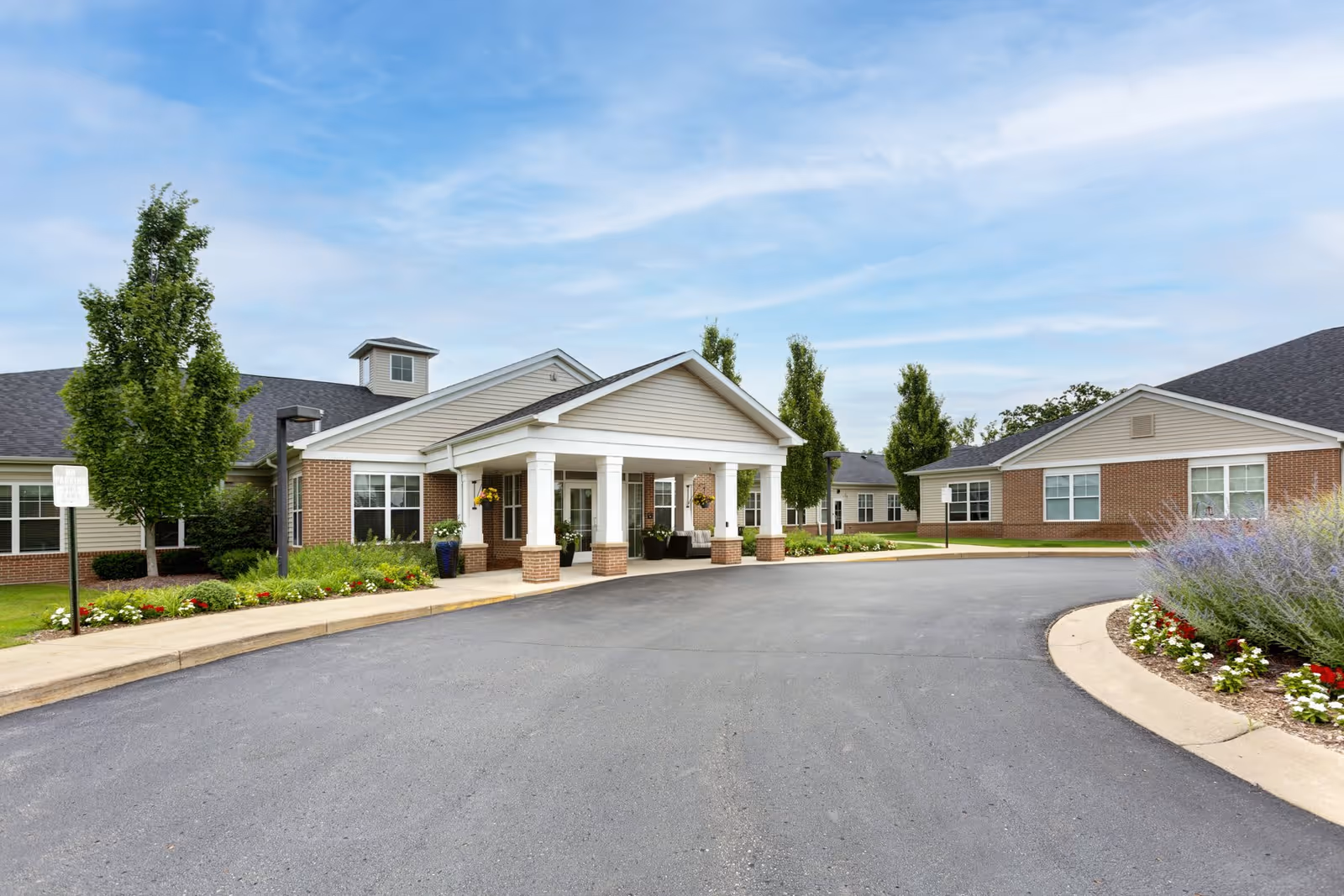 Exterior view of a single-story senior living facility building with a covered entrance supported by white columns. The building has brick and siding walls, surrounded by landscaped greenery, flowers, and trees under a partly cloudy blue sky.