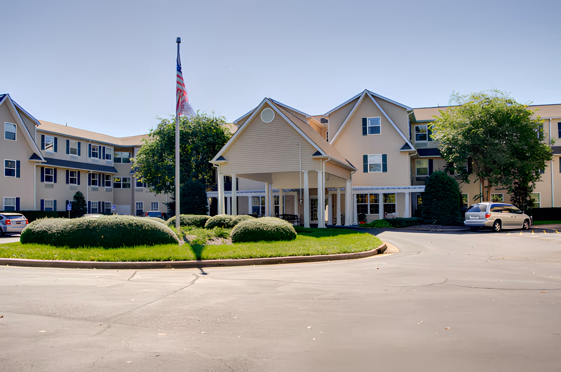 Front exterior view of a large, beige assisted living facility building with multiple windows and a covered entrance supported by white columns. There is a flagpole with an American flag in front, surrounded by neatly trimmed bushes and green grass. Several cars are parked near the building under a clear blue sky.