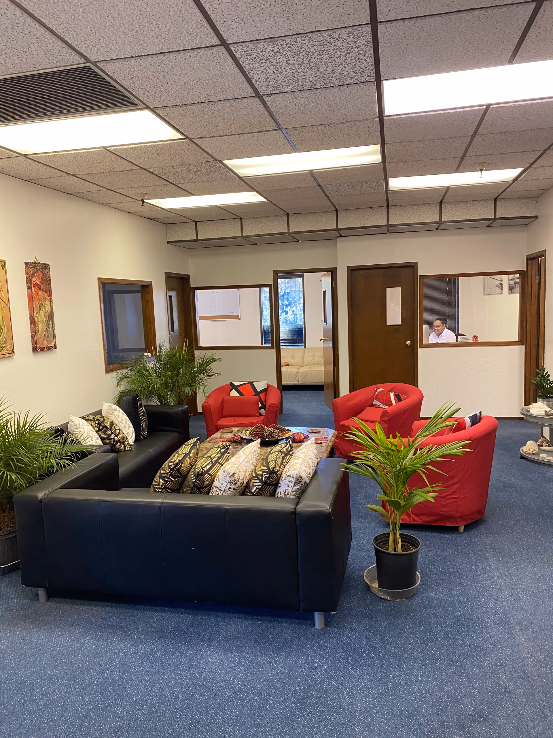 A senior living facility lounge with a black sofa, red chairs, plants, and windows.