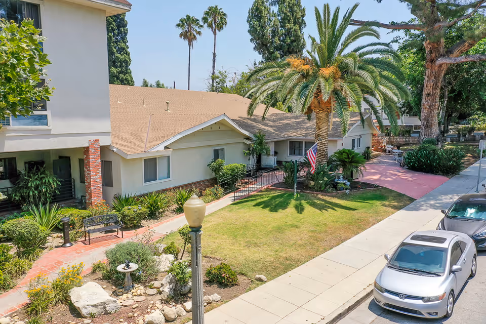Front exterior of a single-story assisted living building with a lawn, palm tree, walkway and parked cars along the street.