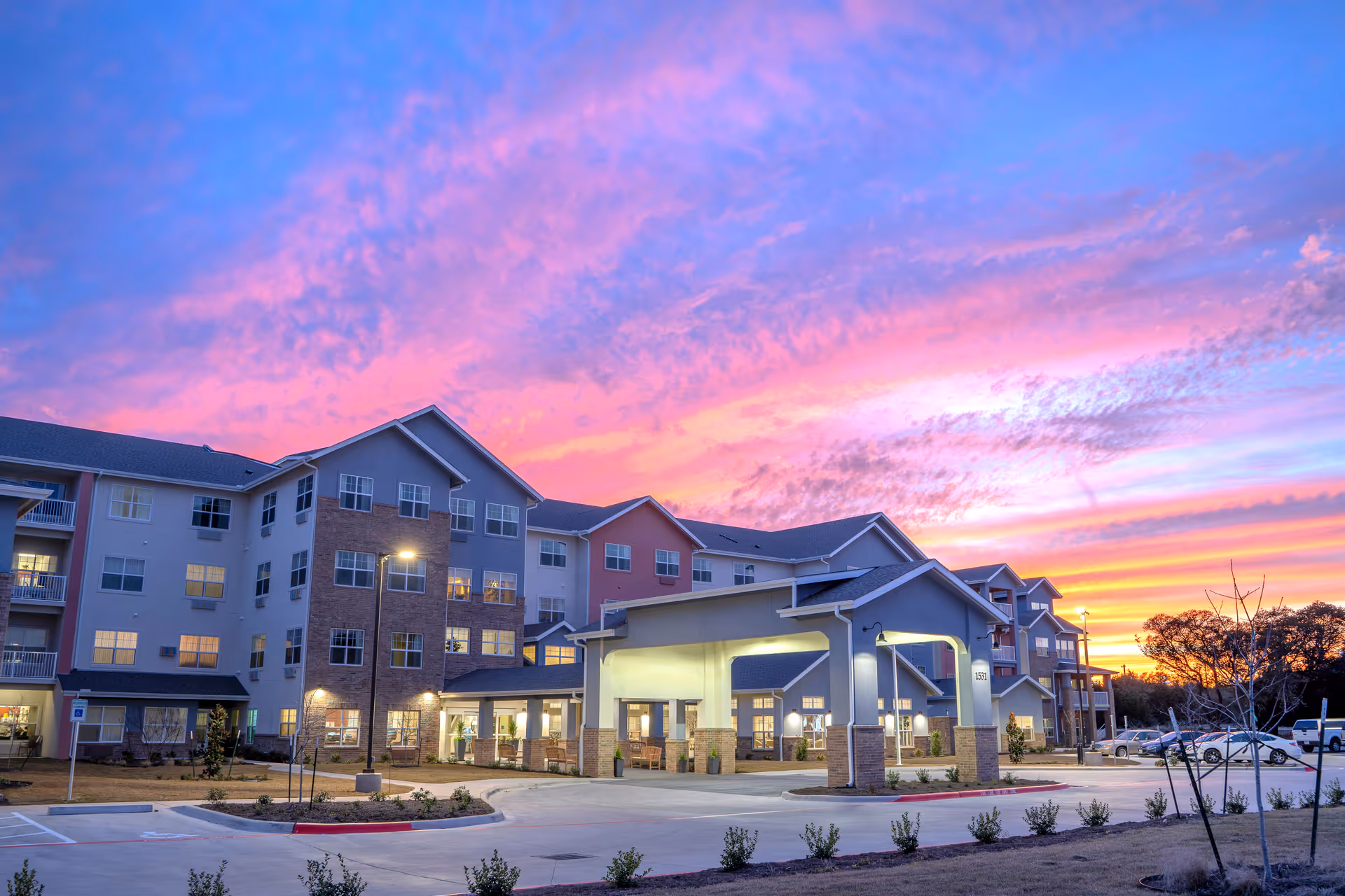 Exterior view of a multi-story senior living facility named Maple Ridge Gracious Retirement Living during sunset, with a colorful sky of pink, purple, and orange hues. The building has a covered entrance with lights on, a parking area, and landscaped surroundings.