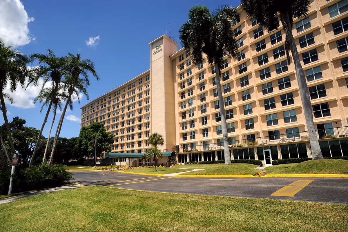 Exterior view of a large multi-story senior living facility building with numerous windows, palm trees, and a well-maintained lawn under a blue sky with some clouds.