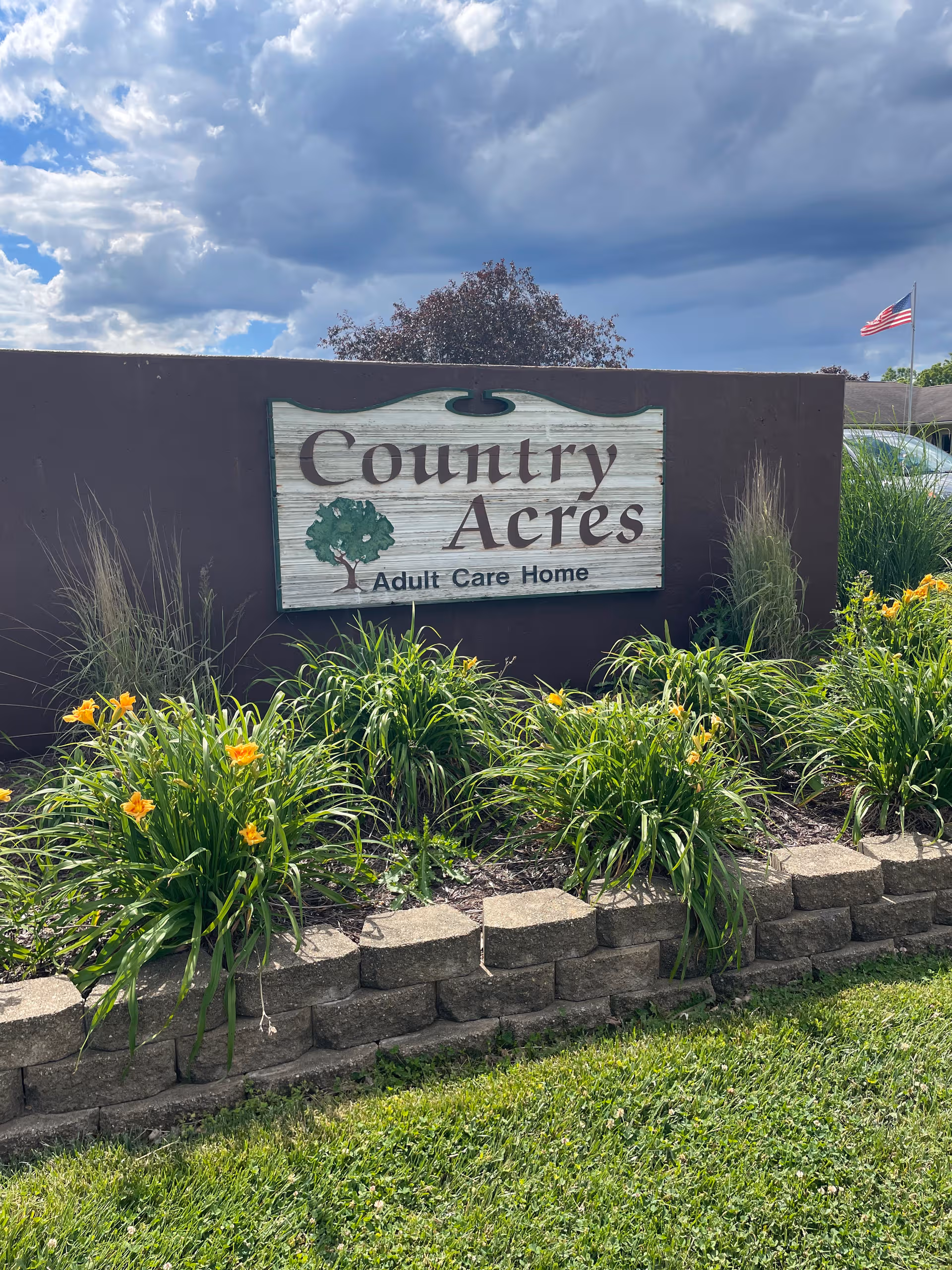 A Country Acres Adult Care Home sign mounted on a dark wall, surrounded by landscaped flowers.