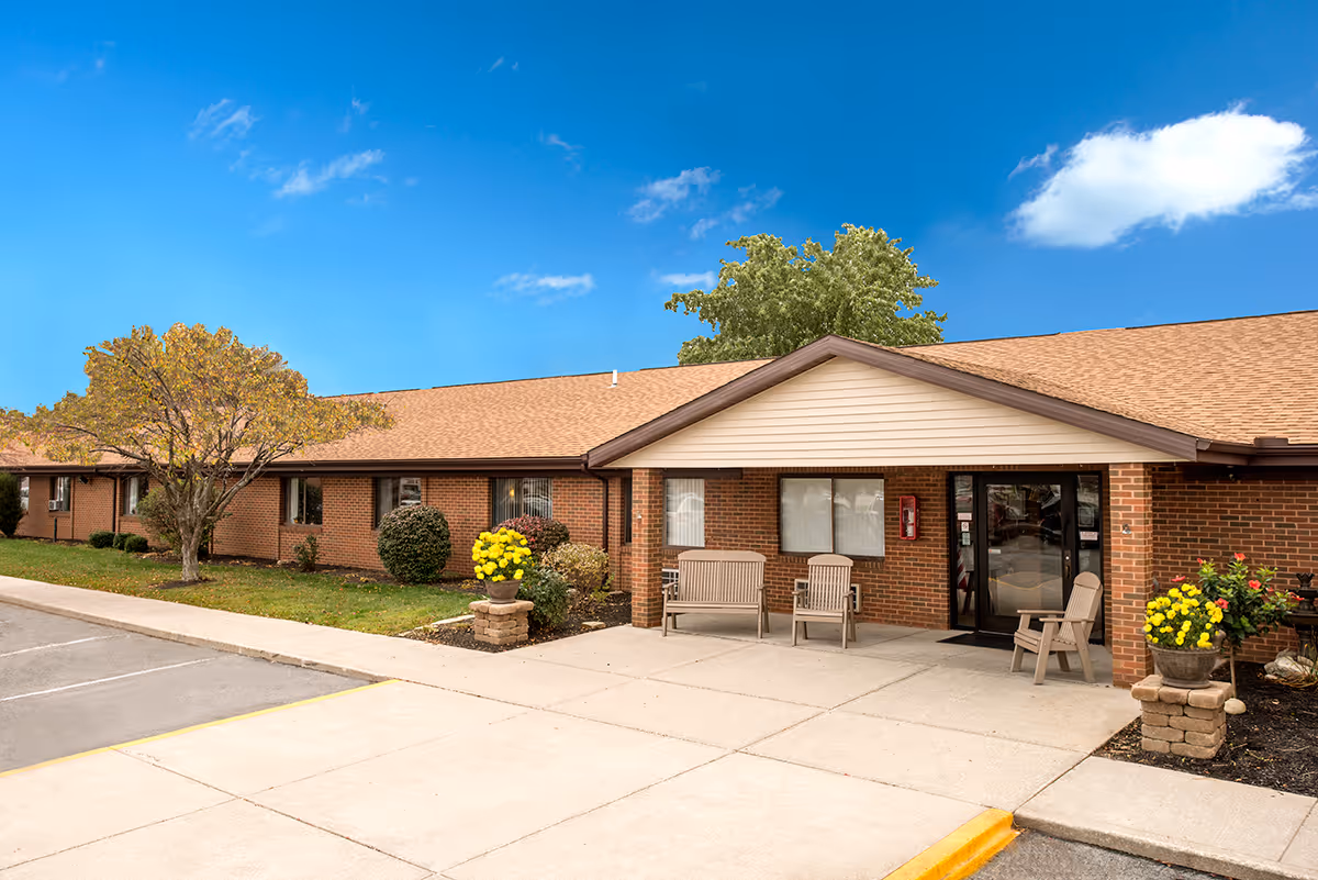 Exterior view of Grande Lake Healthcare Center showing a single-story brick building with a tan roof, a covered entrance with benches and chairs, landscaped bushes and flowers, and a clear blue sky with a few clouds.