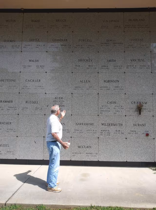 Older man stands before a large outdoor memorial wall engraved with names.