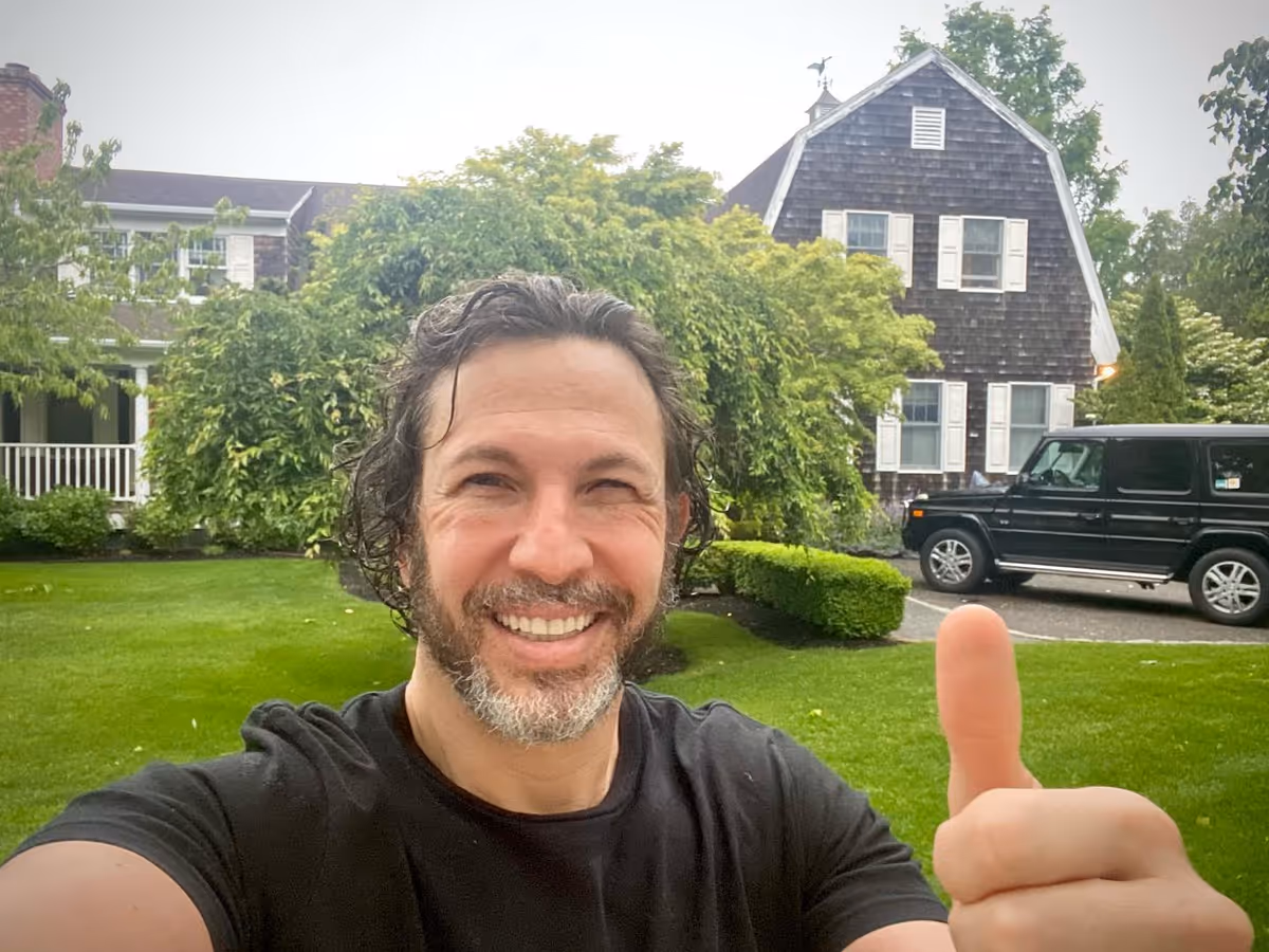 Smiling man takes a selfie outdoors in front of a house-like senior living facility.