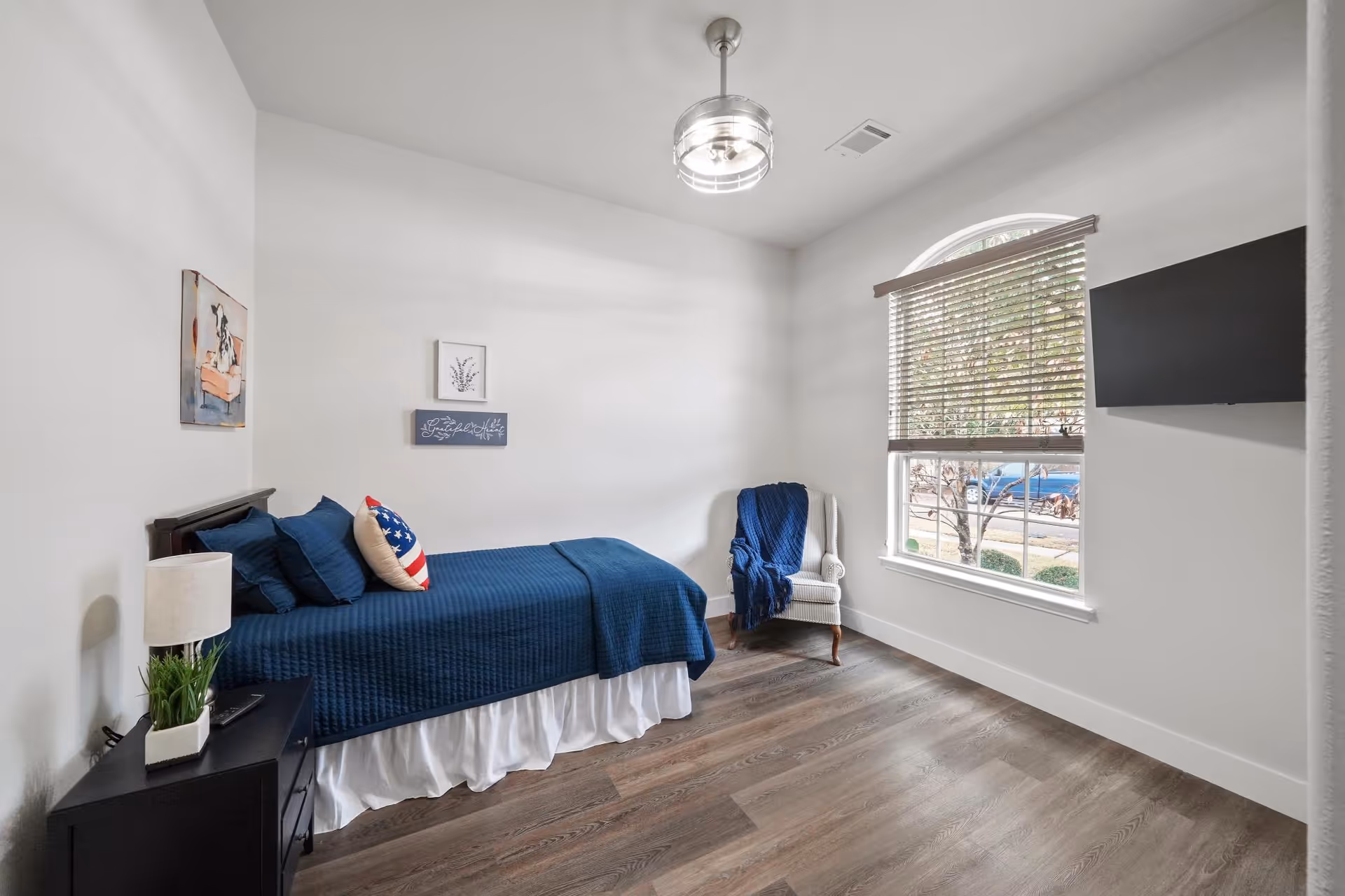 Bedroom in a senior living facility with blue bedding, chair by window, and TV.