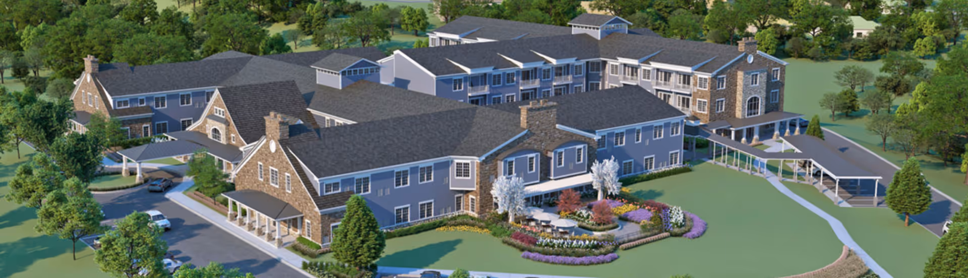 Aerial view of Artisan at Hudson, a large senior living facility with multiple connected buildings featuring gray roofs and stone accents. The grounds include landscaped gardens with colorful flowers, trees, and outdoor seating areas. There is a driveway with parked cars and covered walkways connecting parts of the building.
