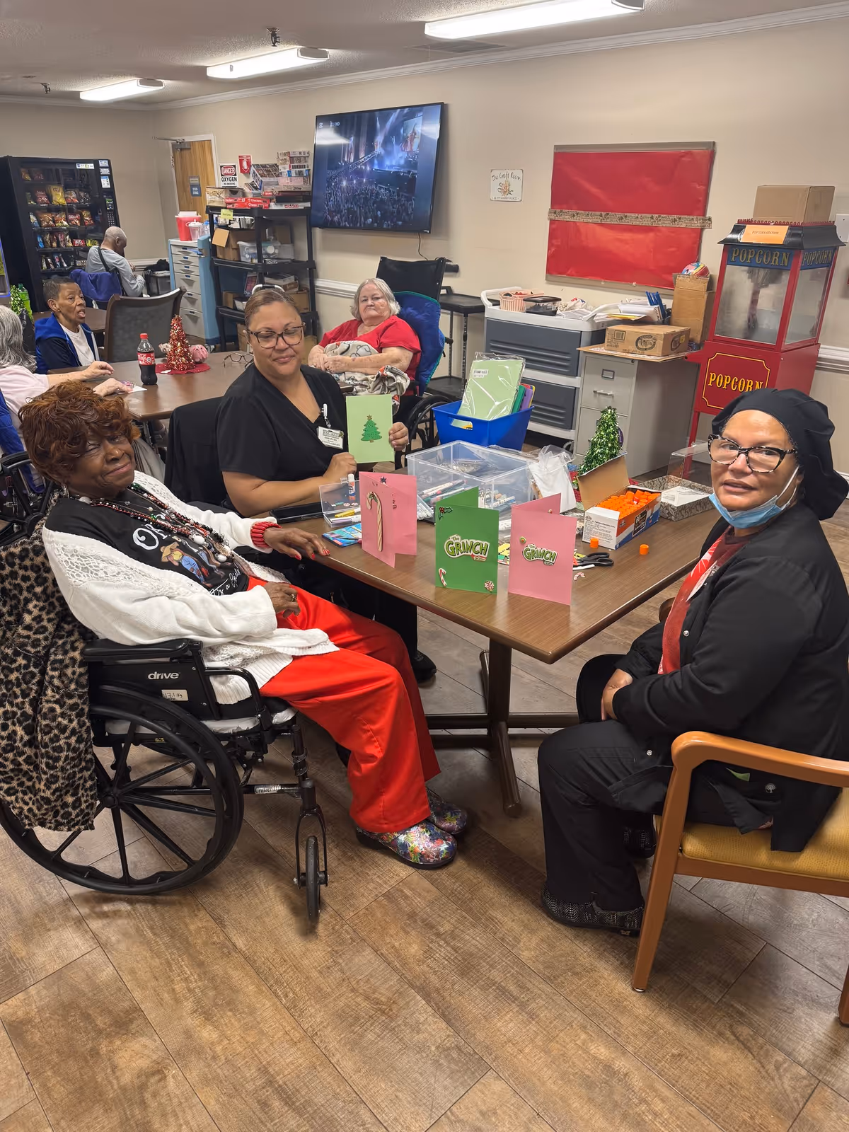 A group of elderly women and a caregiver sitting around a table in a common room at Rocky Mount Rehabilitation Center. They are engaged in a craft activity, making holiday cards. The room has a popcorn machine, a TV mounted on the wall, and various supplies on shelves and tables. Some women are in wheelchairs, and the atmosphere appears warm and social.