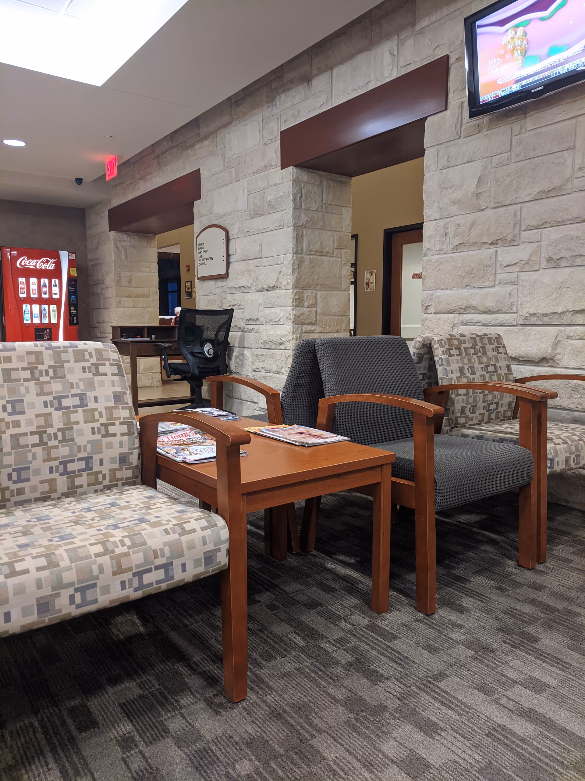A seating area with patterned chairs, wooden tables, and a Coca-Cola vending machine in a hallway.