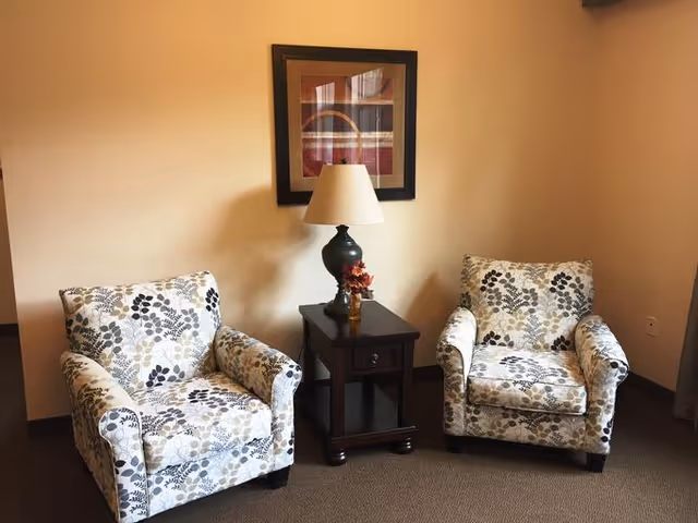 Two patterned armchairs with floral designs are positioned on either side of a dark wooden side table with a drawer and a lower shelf. A lamp with a beige shade and a small decorative item are on the table. A framed abstract artwork hangs on the beige wall behind the table and chairs.