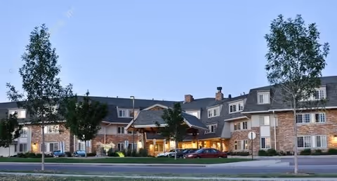 Front exterior of a multi-story brick senior living facility with a covered entry, parked cars, and landscaped grounds.