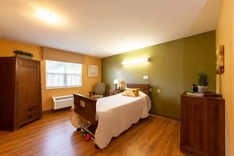 A well-lit bedroom in a senior living facility featuring a single bed with white bedding and a yellow pillow, a wooden armoire, a wooden dresser with a plant and books on top, a comfortable armchair, a window with blinds, and a green accent wall with a wall-mounted light above the bed.