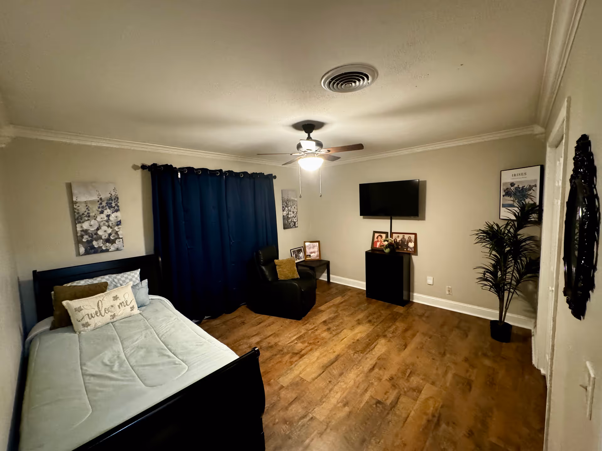 A cozy furnished bedroom with a bed, chair, curtains, wall TV, and wooden floor.