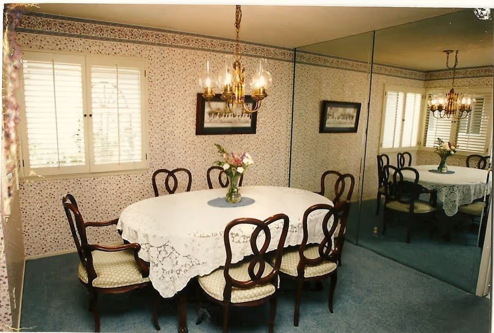 Round dining table with lace tablecloth and six chairs in a floral wallpaper dining room.