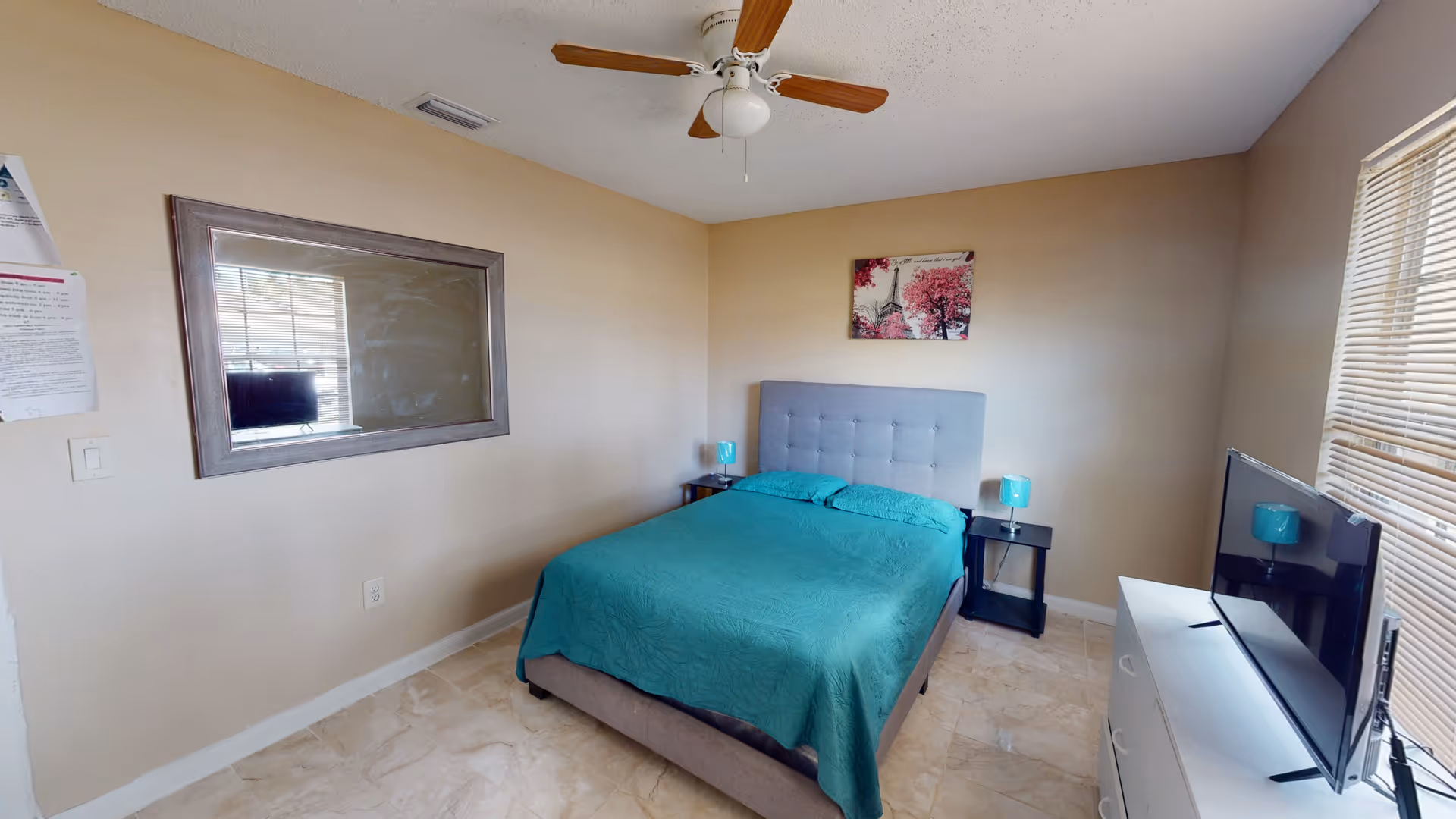 Cozy bedroom with teal bedspread, two nightstands, TV, ceiling fan, and window blinds.