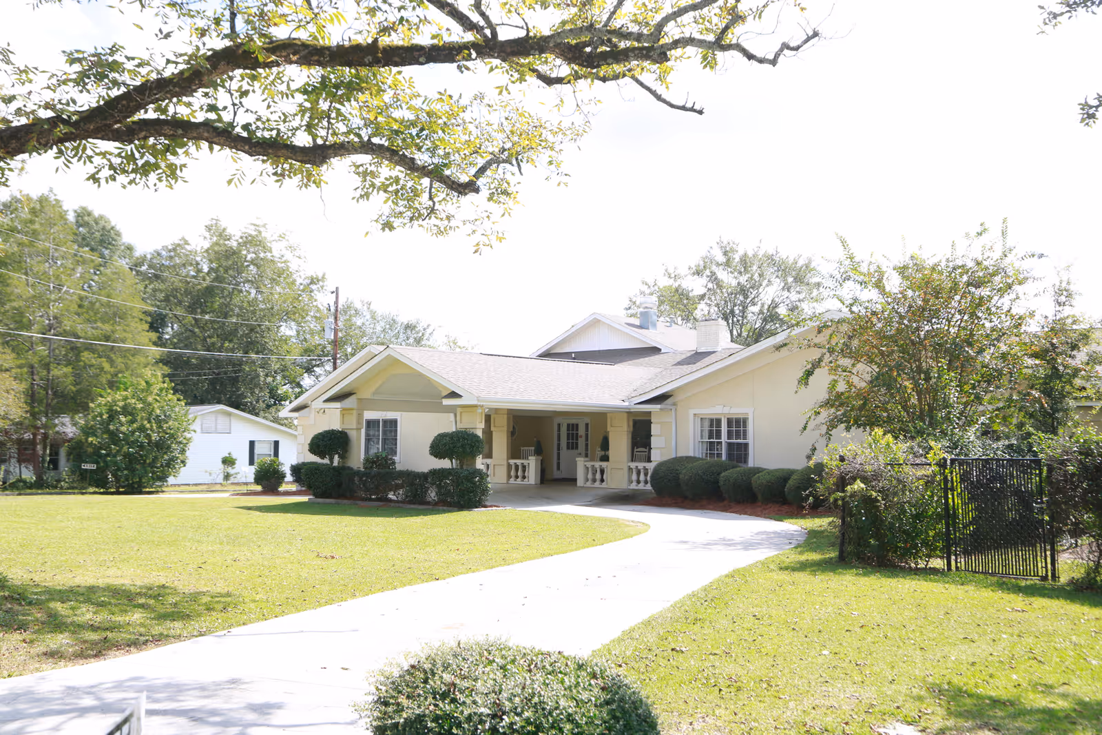 Single-story light-colored building with a covered front porch, curved driveway, and manicured lawn and trees.