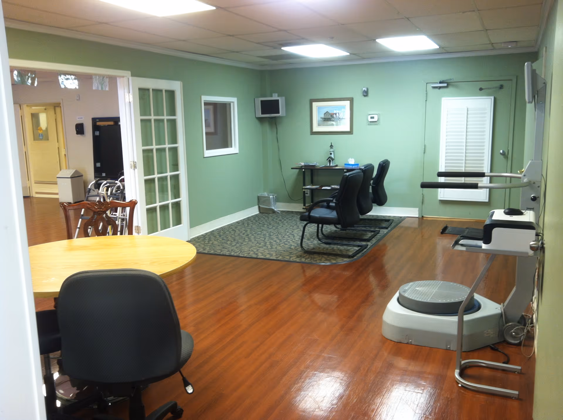 Green-walled common area with round table, chairs, and exercise equipment in a nursing facility.