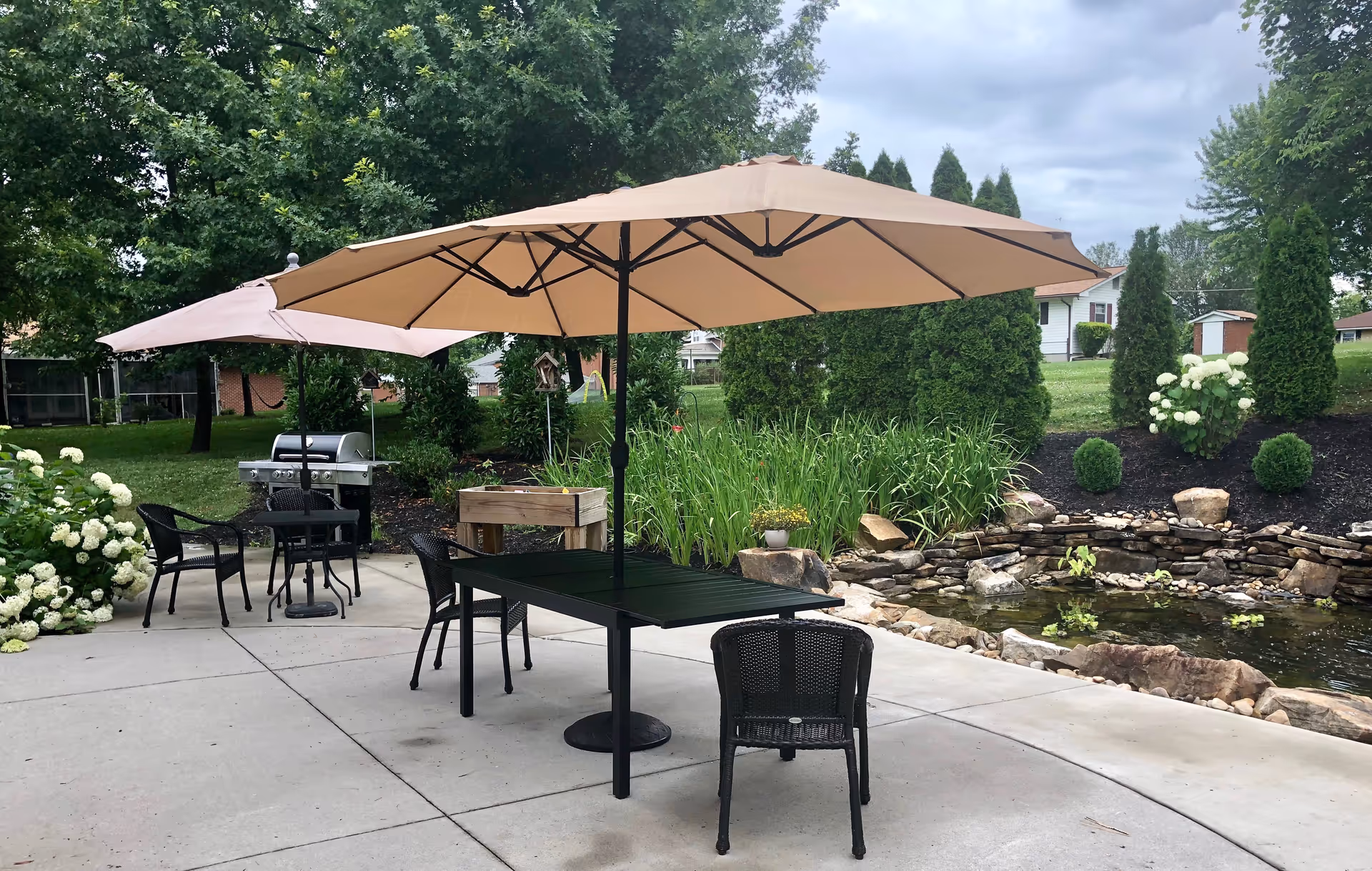 Outdoor patio with beige umbrellas, tables, chairs, a grill, and a small pond.