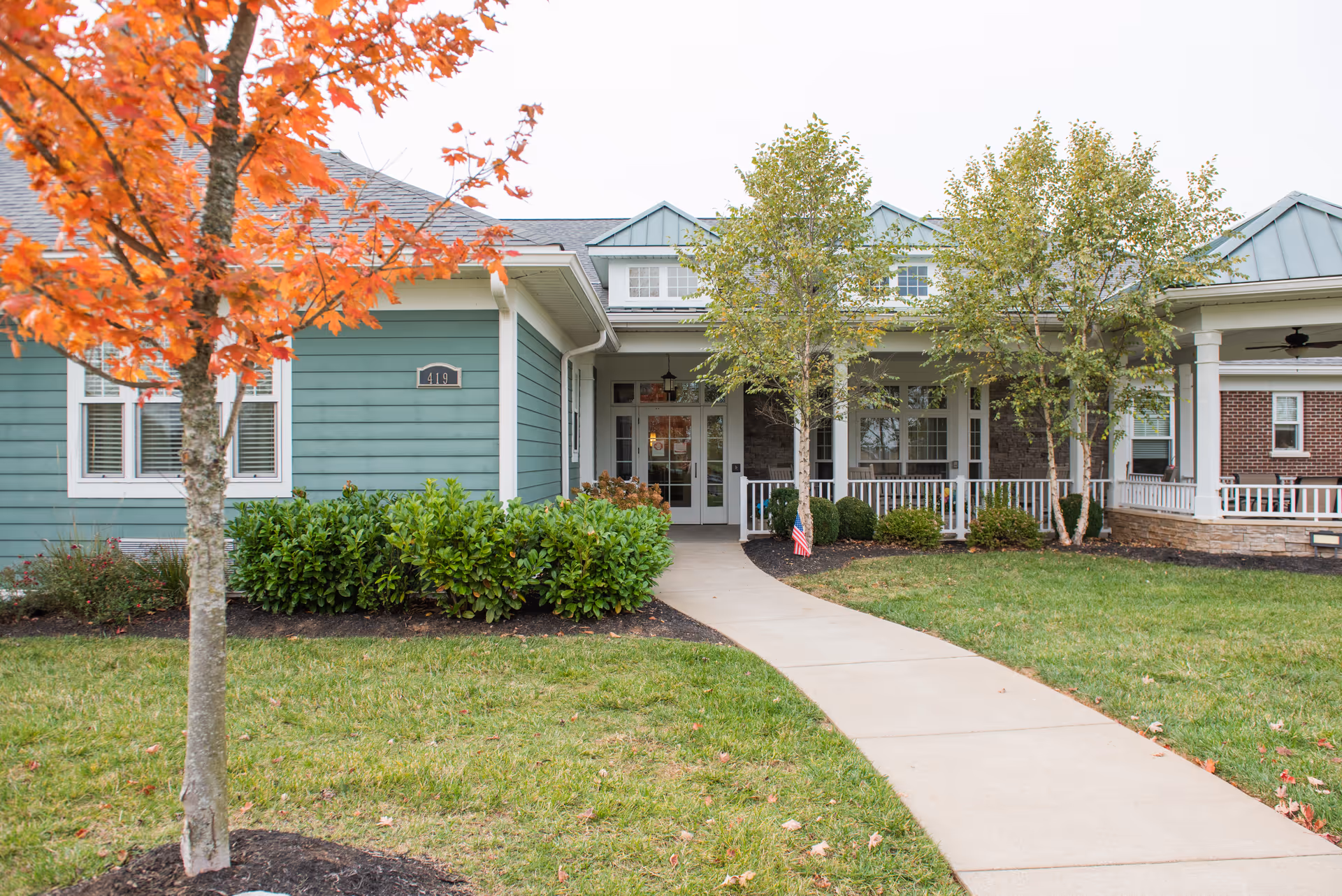 Front exterior view of a senior living facility building with a light blue and brick facade, a concrete walkway leading to the entrance, green bushes, and trees with some autumn-colored leaves.