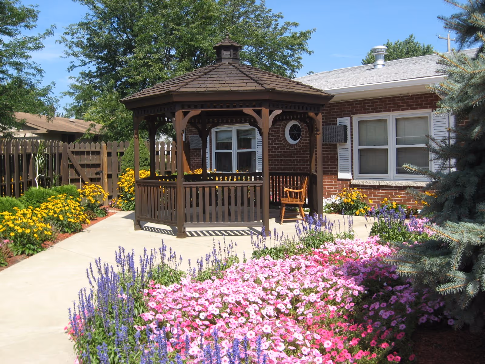 A garden area with a wooden gazebo surrounded by colorful flowers including pink, purple, and yellow blooms. There is a brick building with white window shutters in the background and a wooden fence enclosing the garden.