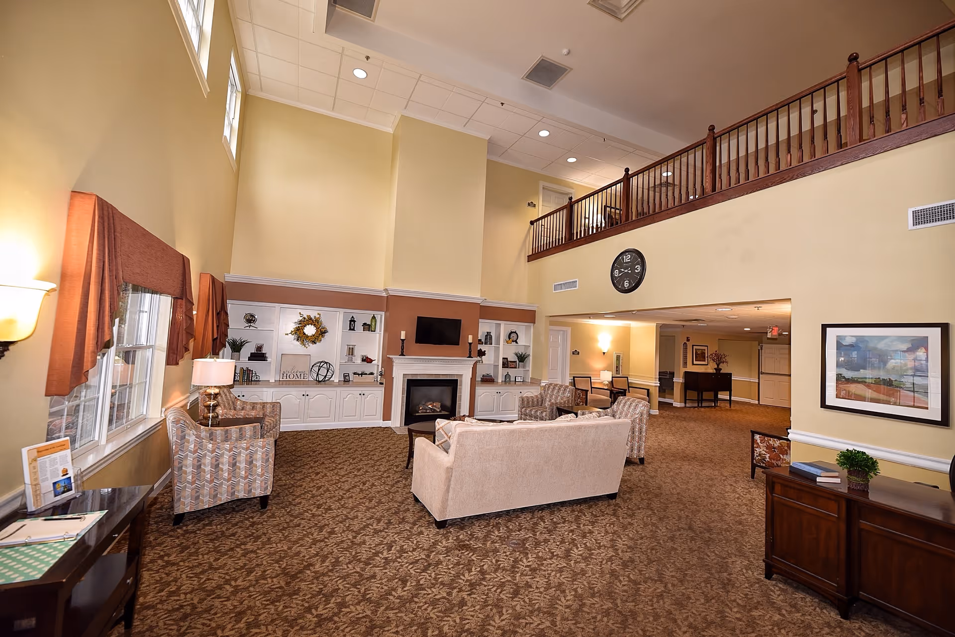 A spacious senior living lobby with sofas, a fireplace, built-in shelves, and a second-floor railing.