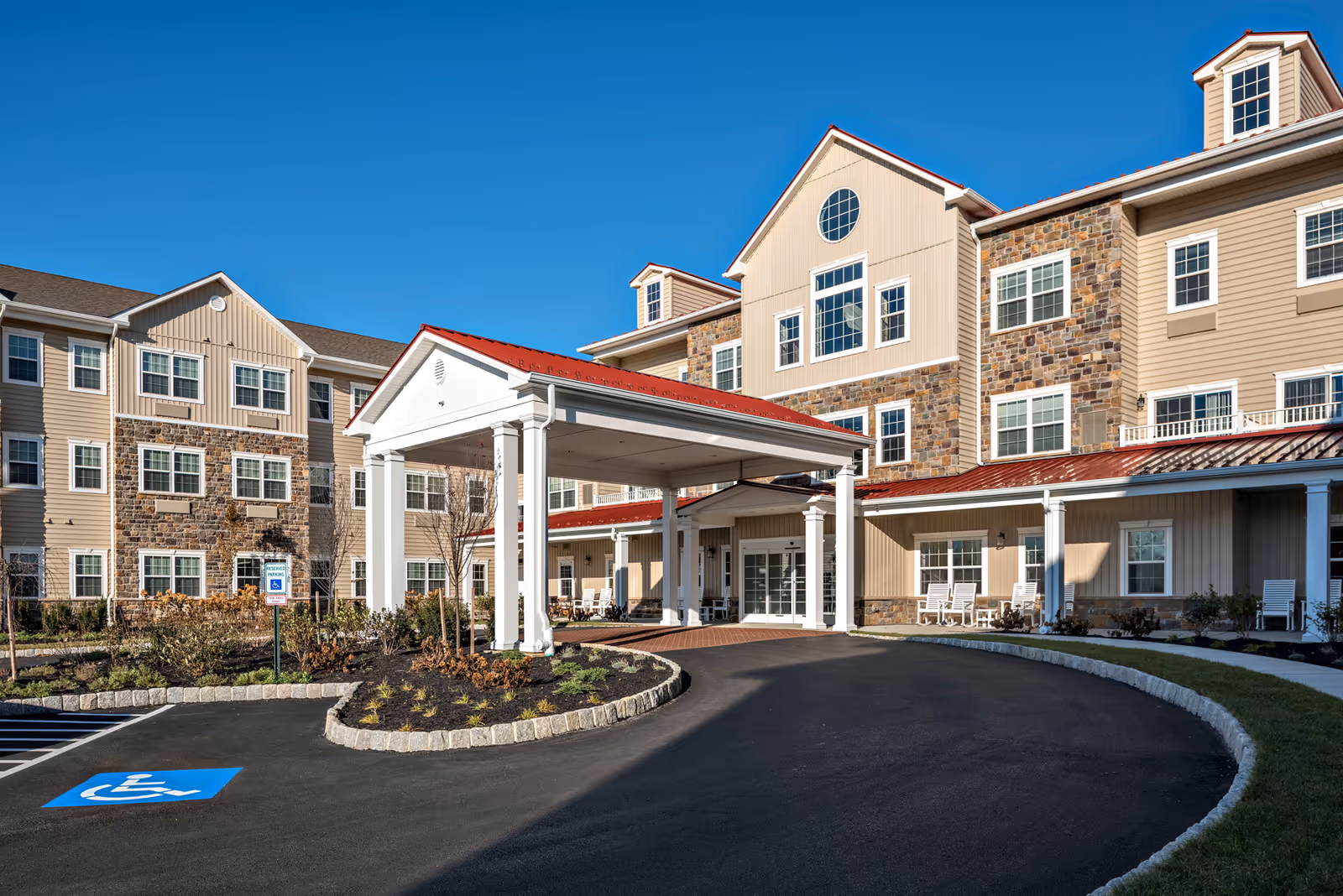 Exterior view of Merrill Gardens at West Chester, showing a multi-story building with beige siding and stone accents. The entrance features a covered driveway with white columns and a red roof. There is a handicapped parking space visible in the foreground, and the sky is clear and blue.