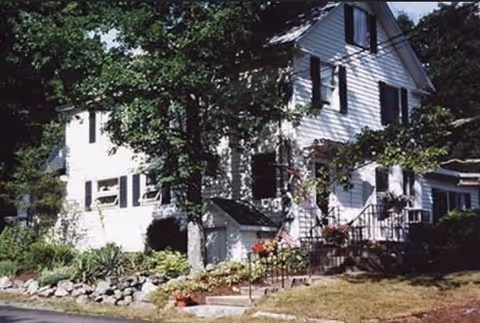 A white two-story house with black shutters surrounded by trees and landscaped garden beds with rocks and plants. There are steps leading up to the front door with a black railing and some potted flowers near the entrance.