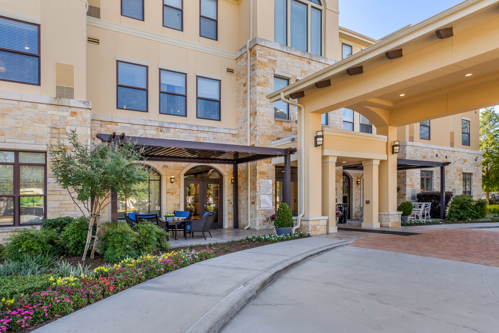 Front entrance of a senior living facility with a covered driveway, stone facade, outdoor seating area, and landscaped flower beds.