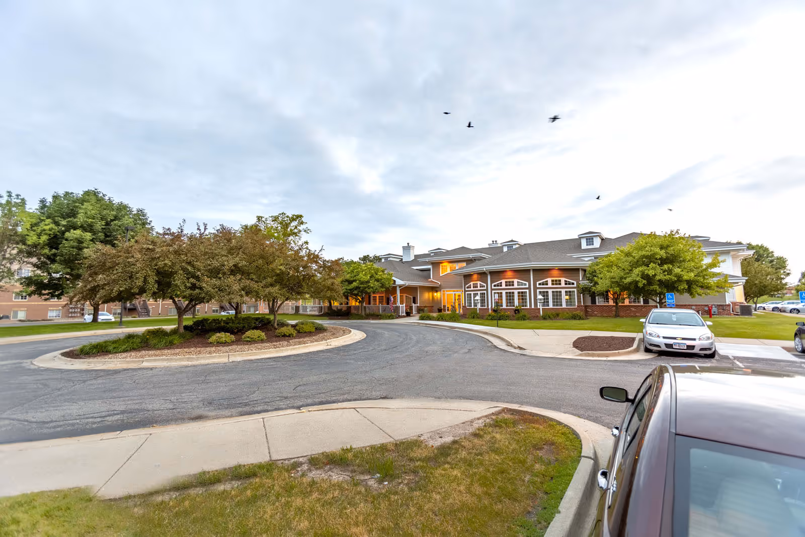 Front exterior of a single-story senior living building with a circular driveway, landscaped islands and parked cars under a cloudy sky.