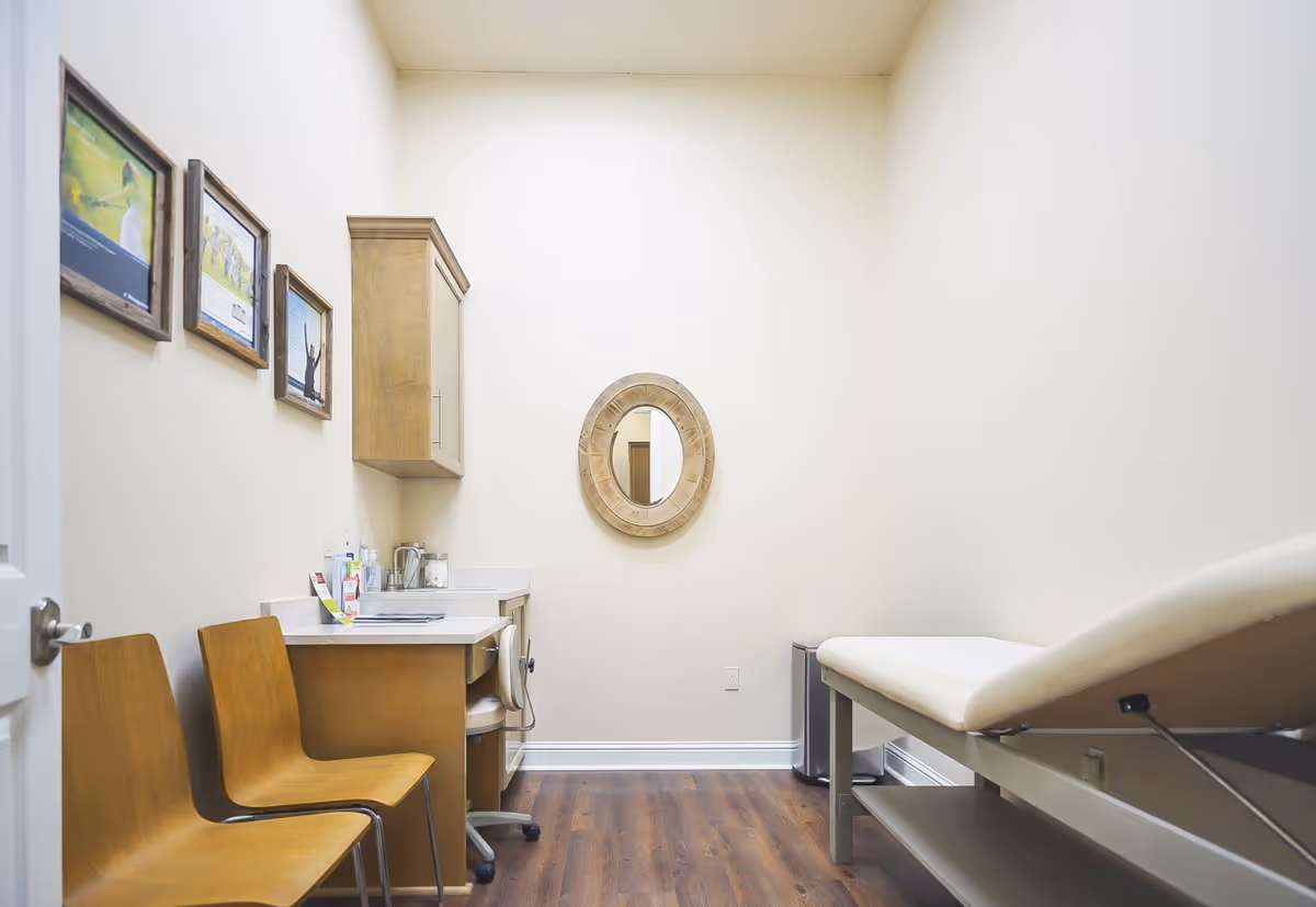 Small clinic exam room with an examination table, chairs, a sink, and a round wall mirror.
