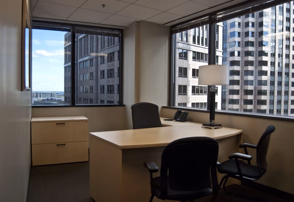 Office workspace with a desk, chairs, and large windows overlooking city skyscrapers.