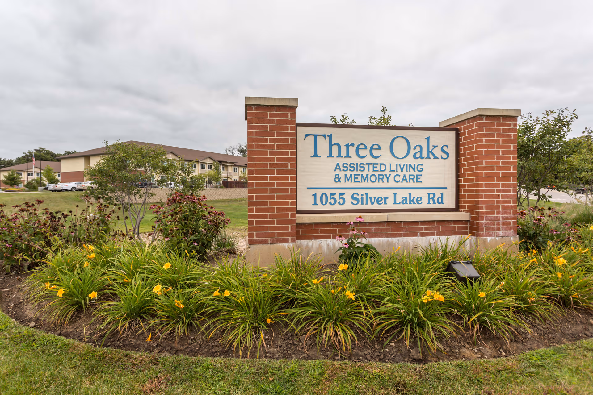 Brick sign for Three Oaks Assisted Living and Memory Care with landscaping and distant building.