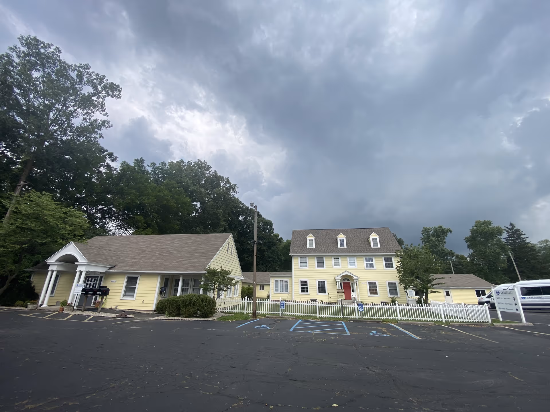 Yellow residential-style facility building with white fencing and a parking lot under cloudy skies.