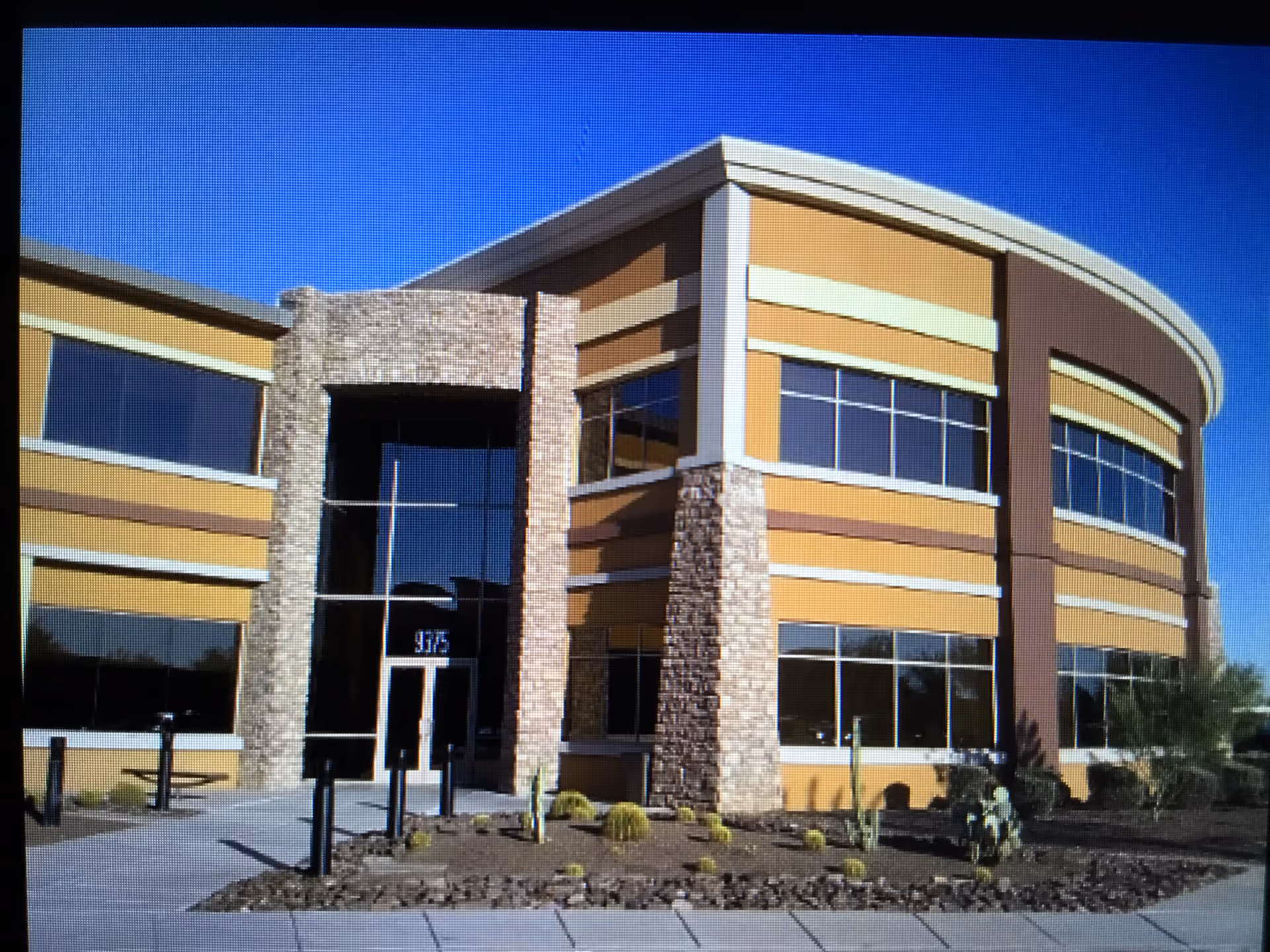 Modern two-story building with stone entry pillars, large windows, and desert landscaping under a blue sky.