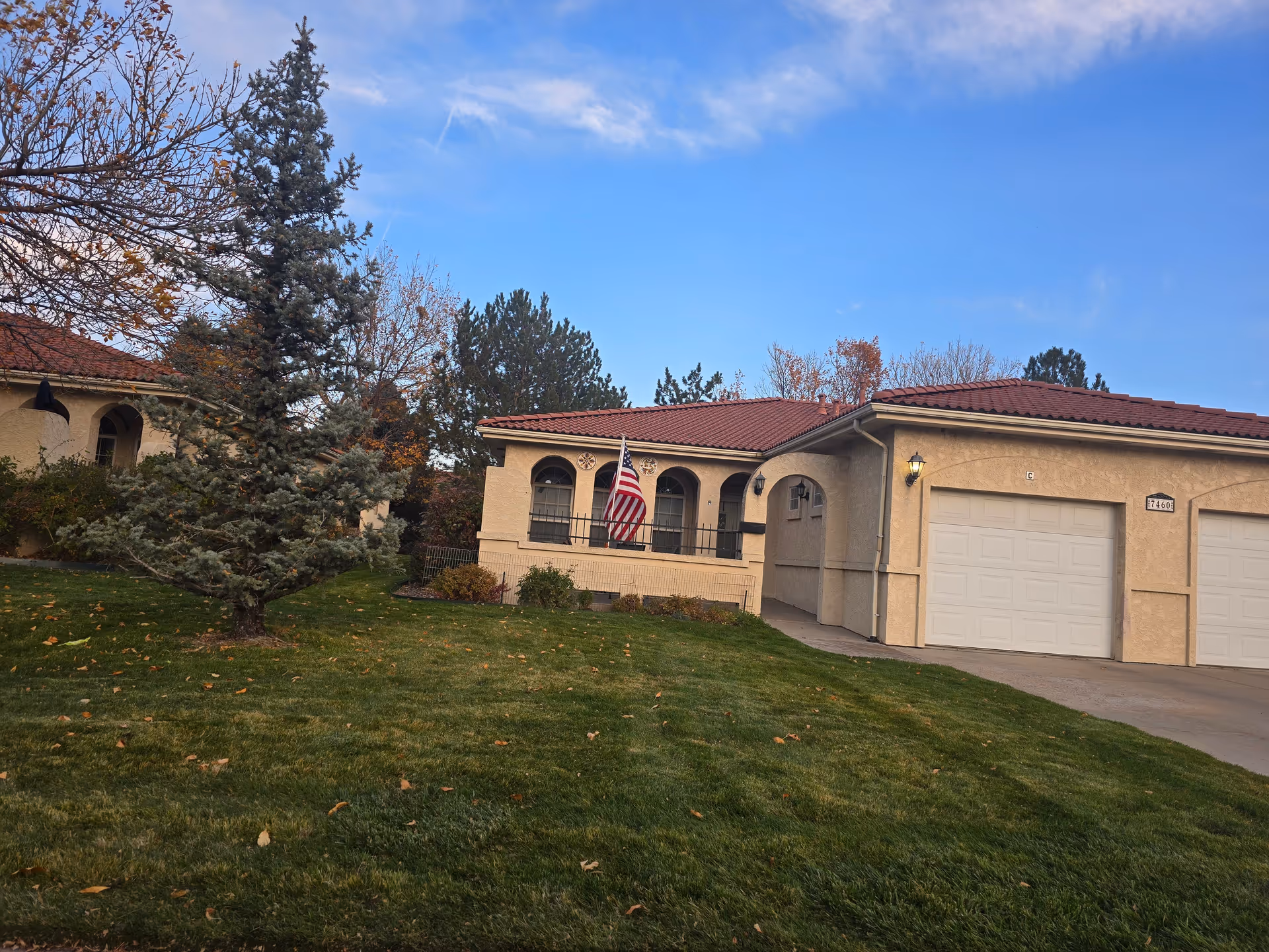 Front exterior of a single-story stucco building with arched windows, an American flag, attached garages, and a grassy lawn.